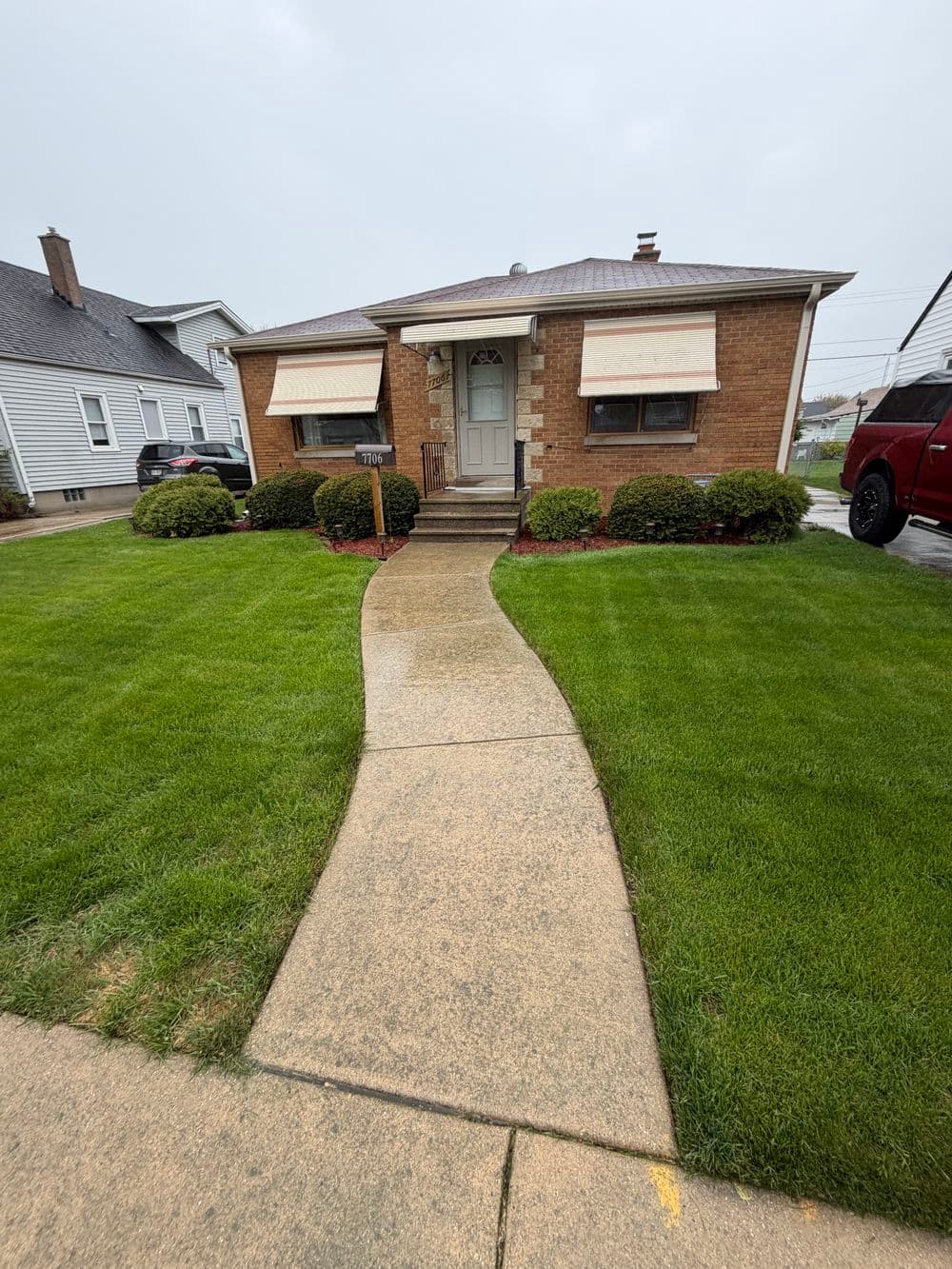 Brick house with awnings, landscaped lawn, and paved walkway on a rainy day.
