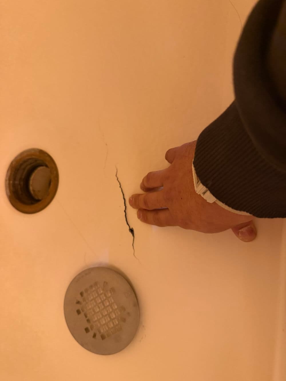 Hand inspecting a crack in a shower tub with drain visible. Home repair concern.
